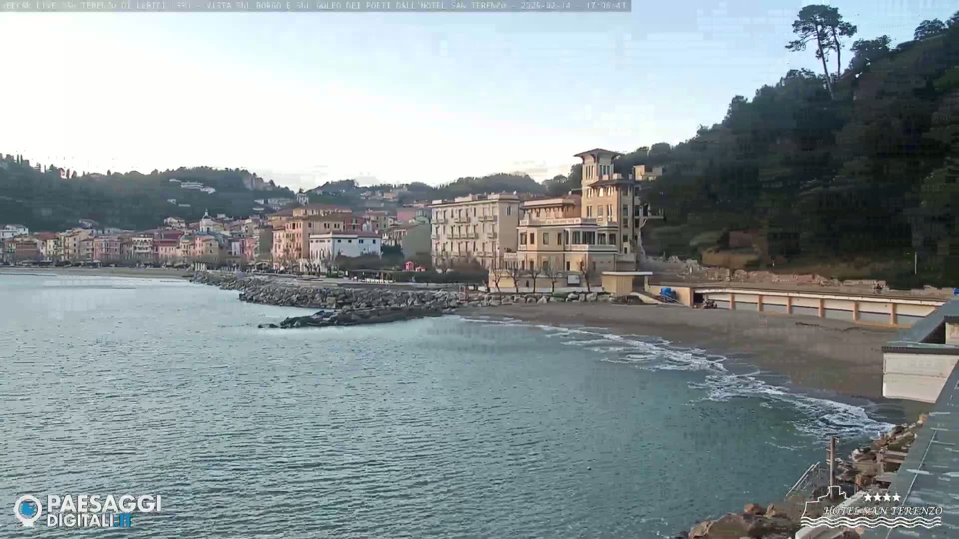 A calm sea laps a sandy beach in front of a collection of buildings, backed by hills under a clear sky.