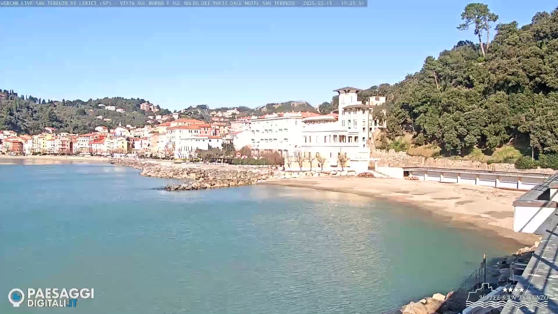 A coastal town with a sandy beach, calm blue water, and buildings nestled against a hillside under a clear sunny sky.