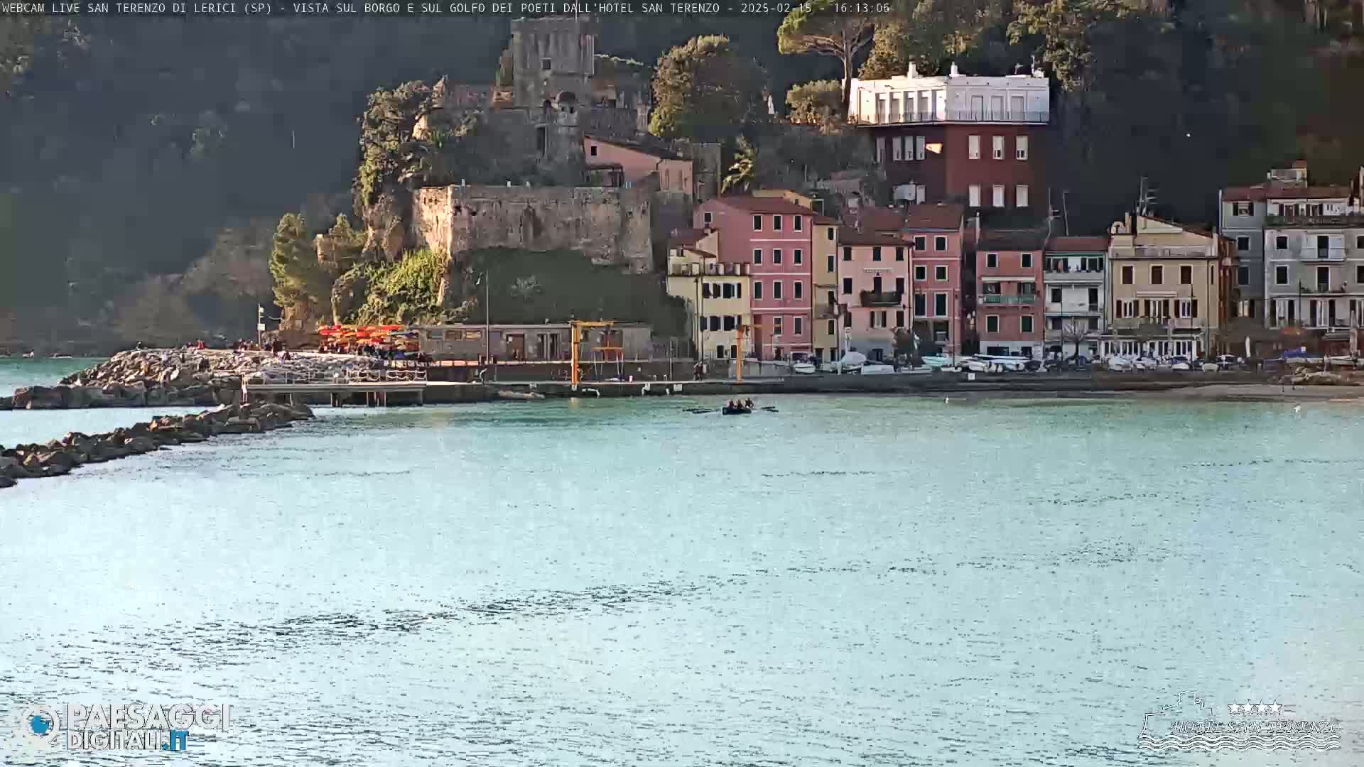 A calm body of water sits in front of a coastal town built into a hillside, under a mostly clear sky.