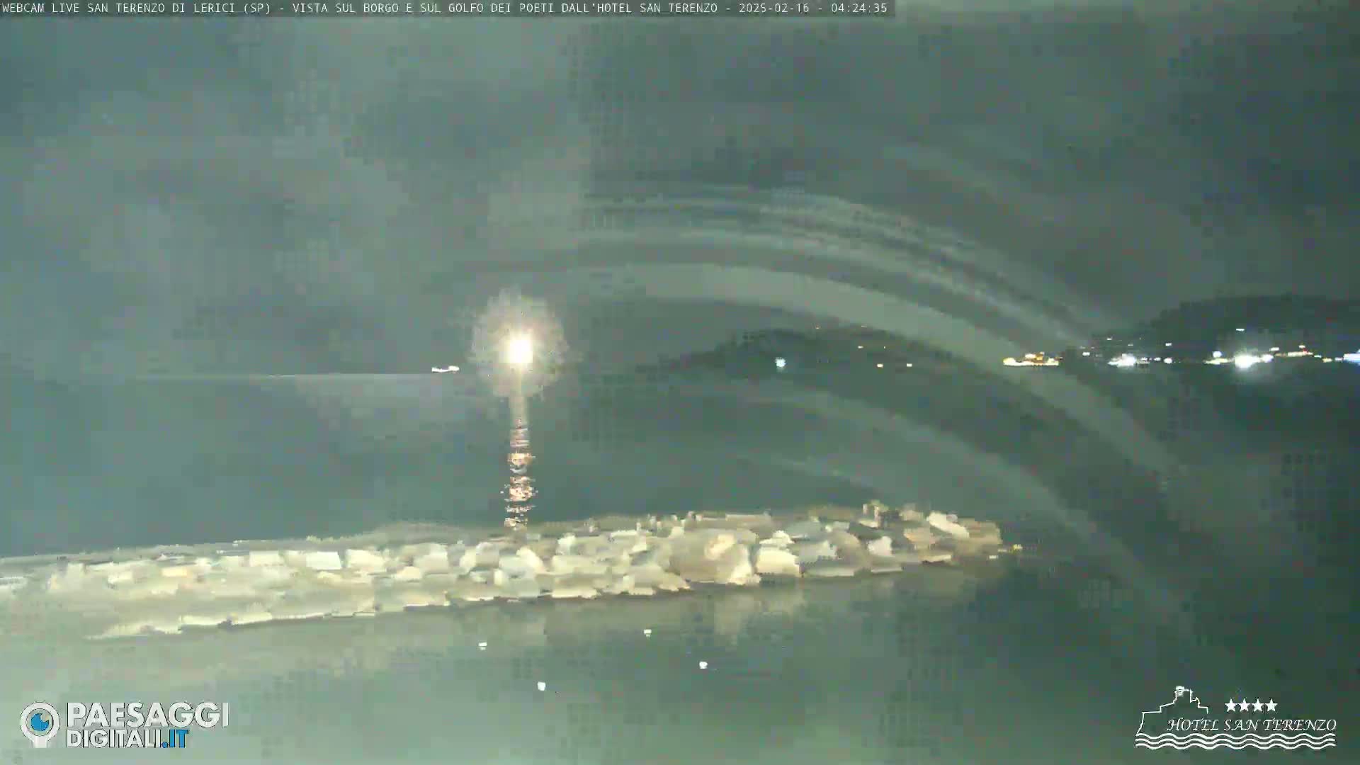 A nighttime view of a calm body of water with a rocky breakwater extending into it, under a dark sky with faint light streaks.
