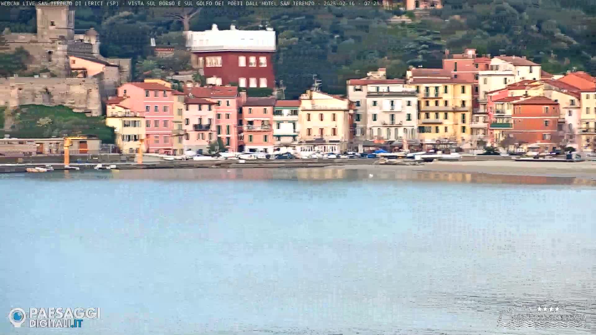 A calm body of water reflects a row of colorful buildings nestled against a hillside.