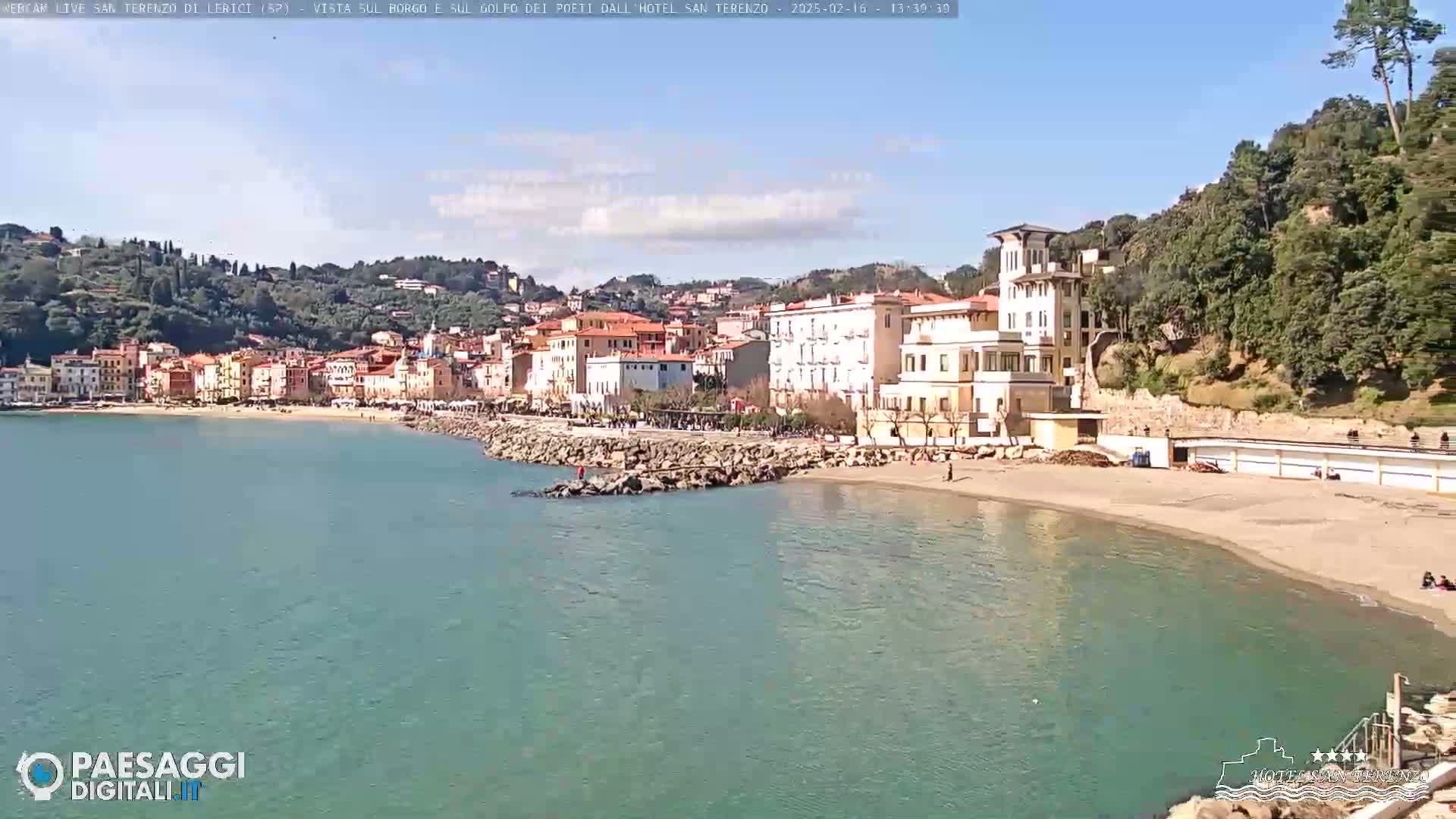 A picturesque coastal town with pastel-colored buildings sits on a calm, teal-colored bay under a mostly sunny sky.