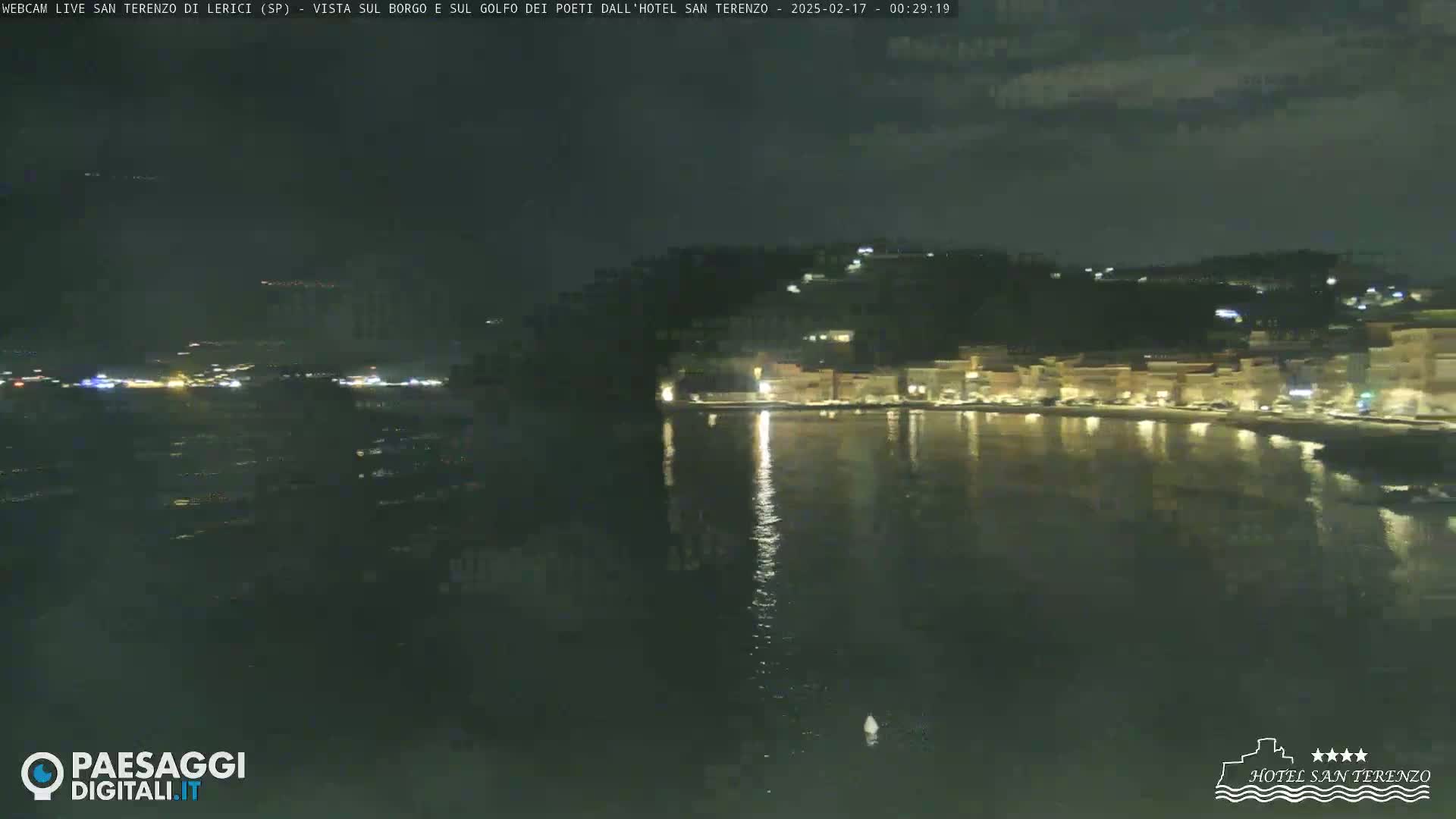 A nighttime view of a waterfront town with lights reflecting on calm water under a partly cloudy sky.