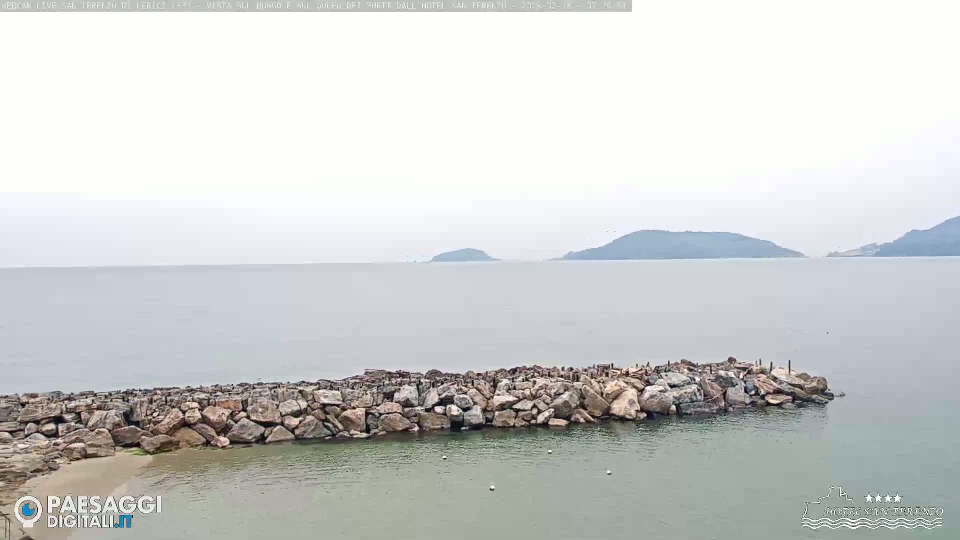 A rocky breakwater protects a calm, light-green bay under an overcast sky, with distant hills visible across the hazy water.