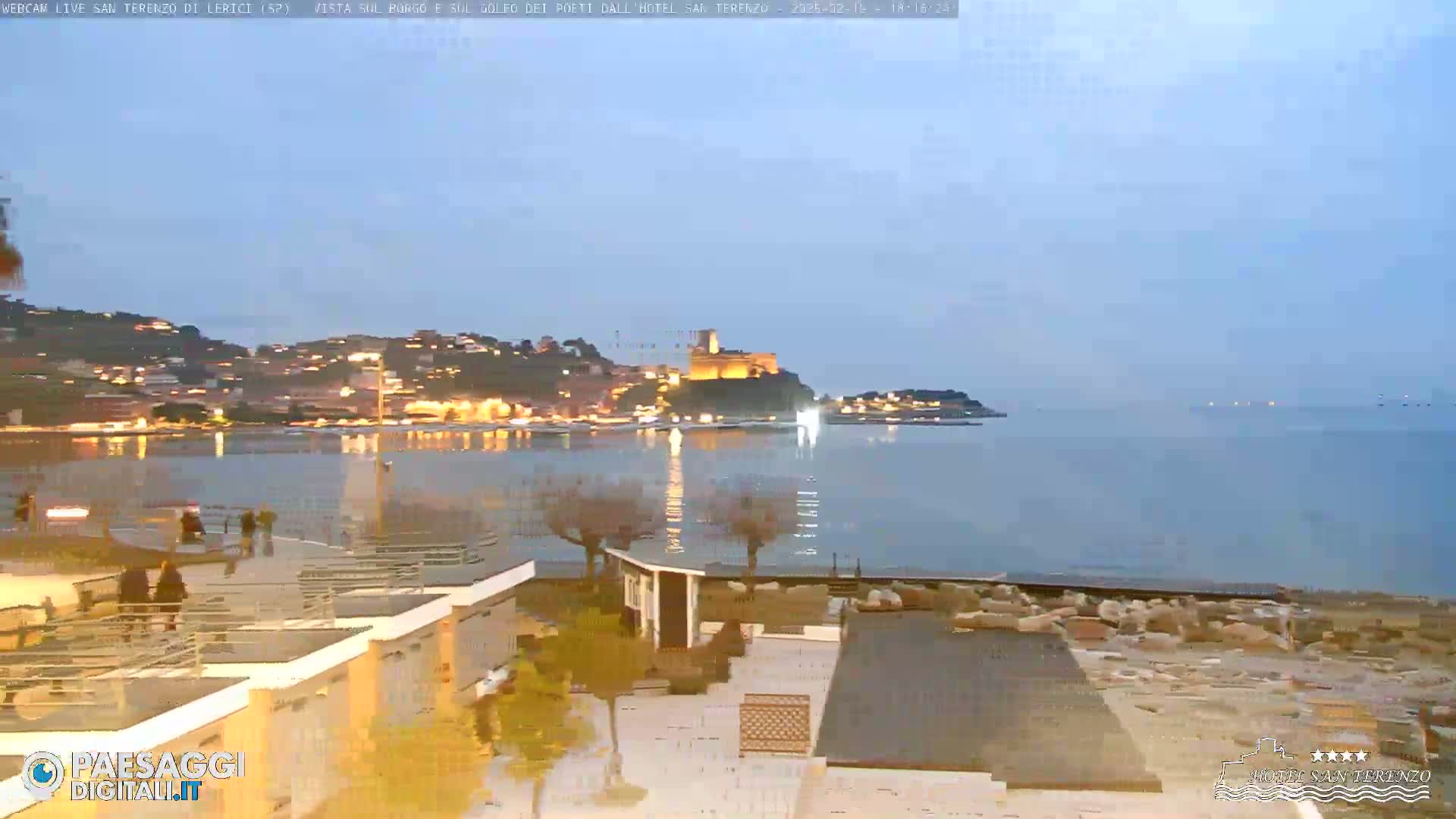 A seaside town at dusk, with lights twinkling along the shore and a castle perched on a hill overlooking a calm body of water.