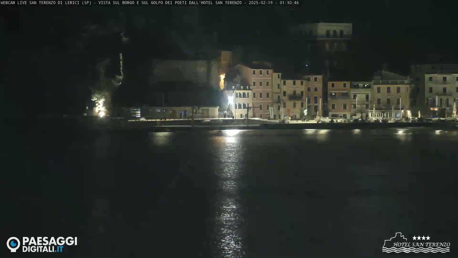 A nighttime view of a waterfront town with buildings illuminated along the shore, reflecting in calm water.