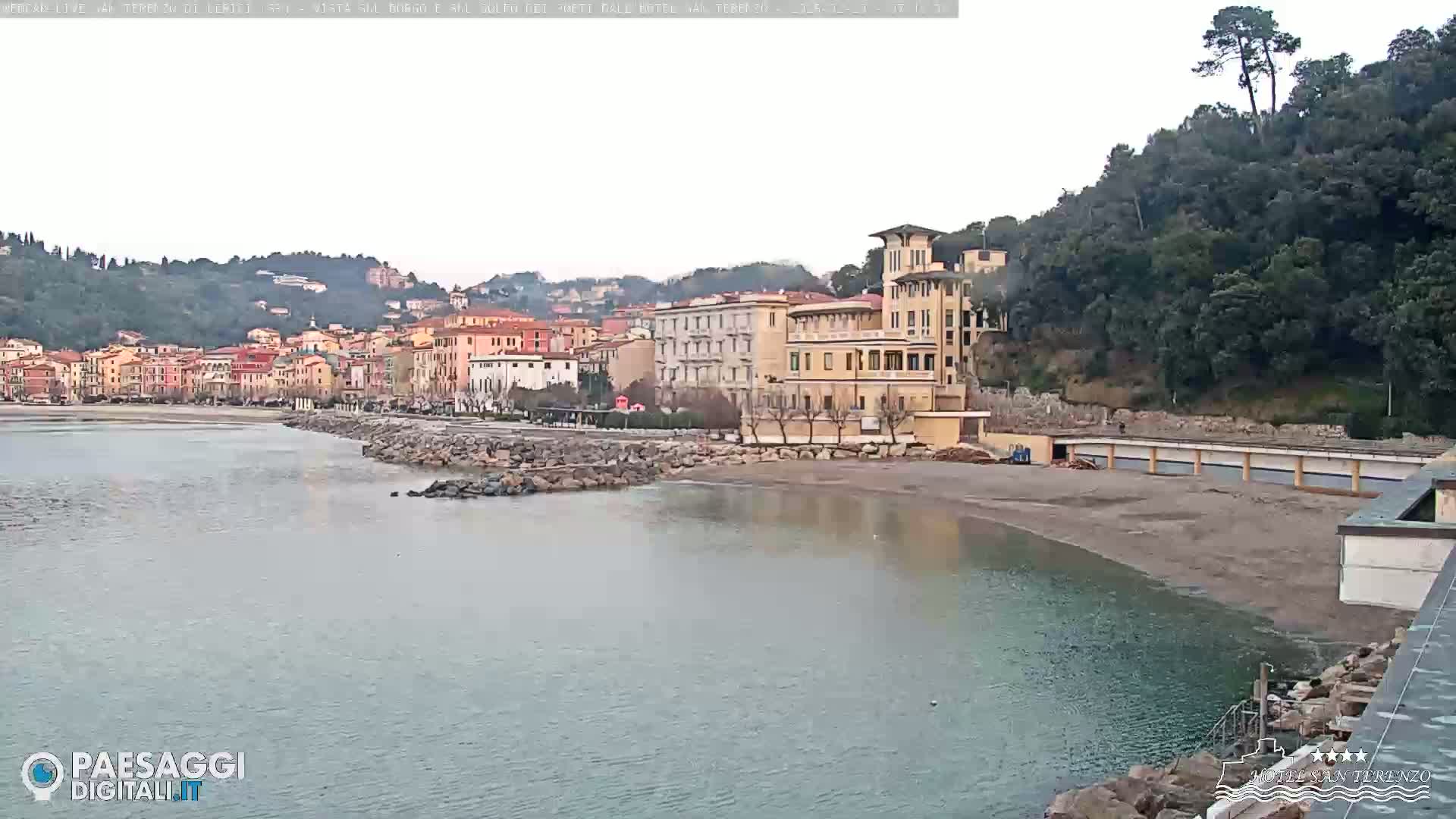 A coastal town with multi-story buildings sits on a hill overlooking a calm bay under an overcast sky.