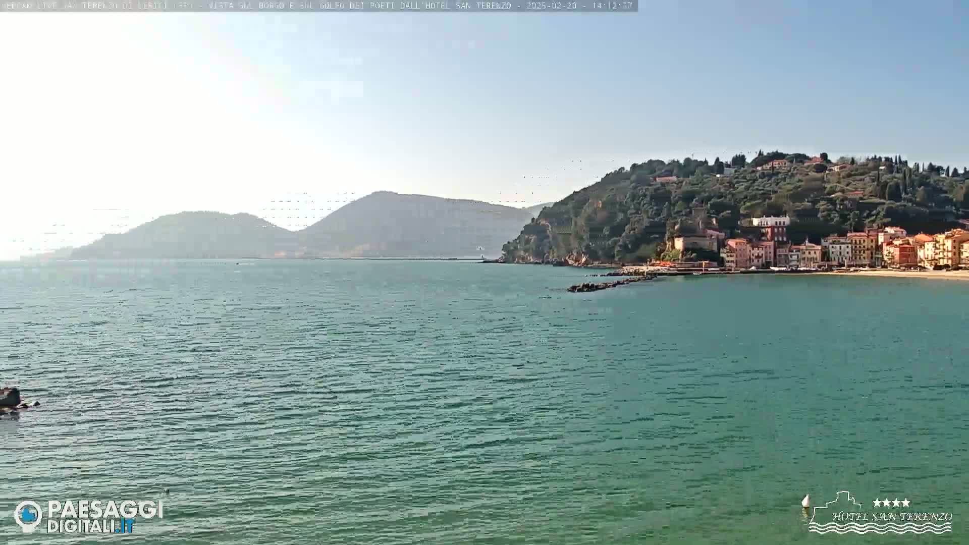 A picturesque coastal town nestled on a hillside overlooking a calm, teal sea under a clear sky.