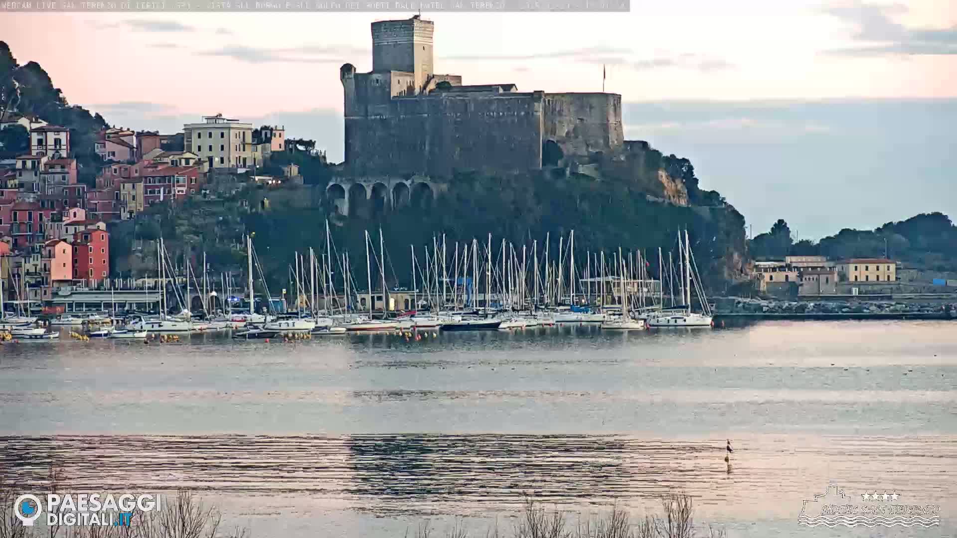 A stone castle sits atop a hill overlooking a harbor filled with sailboats, under a mostly clear sky.