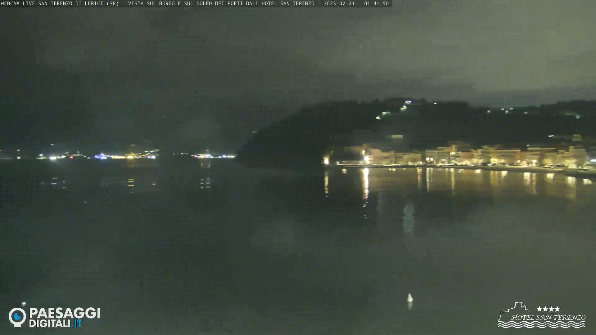 A nighttime view of a calm body of water reflecting the lights of a town nestled on a hillside under a dark, overcast sky.