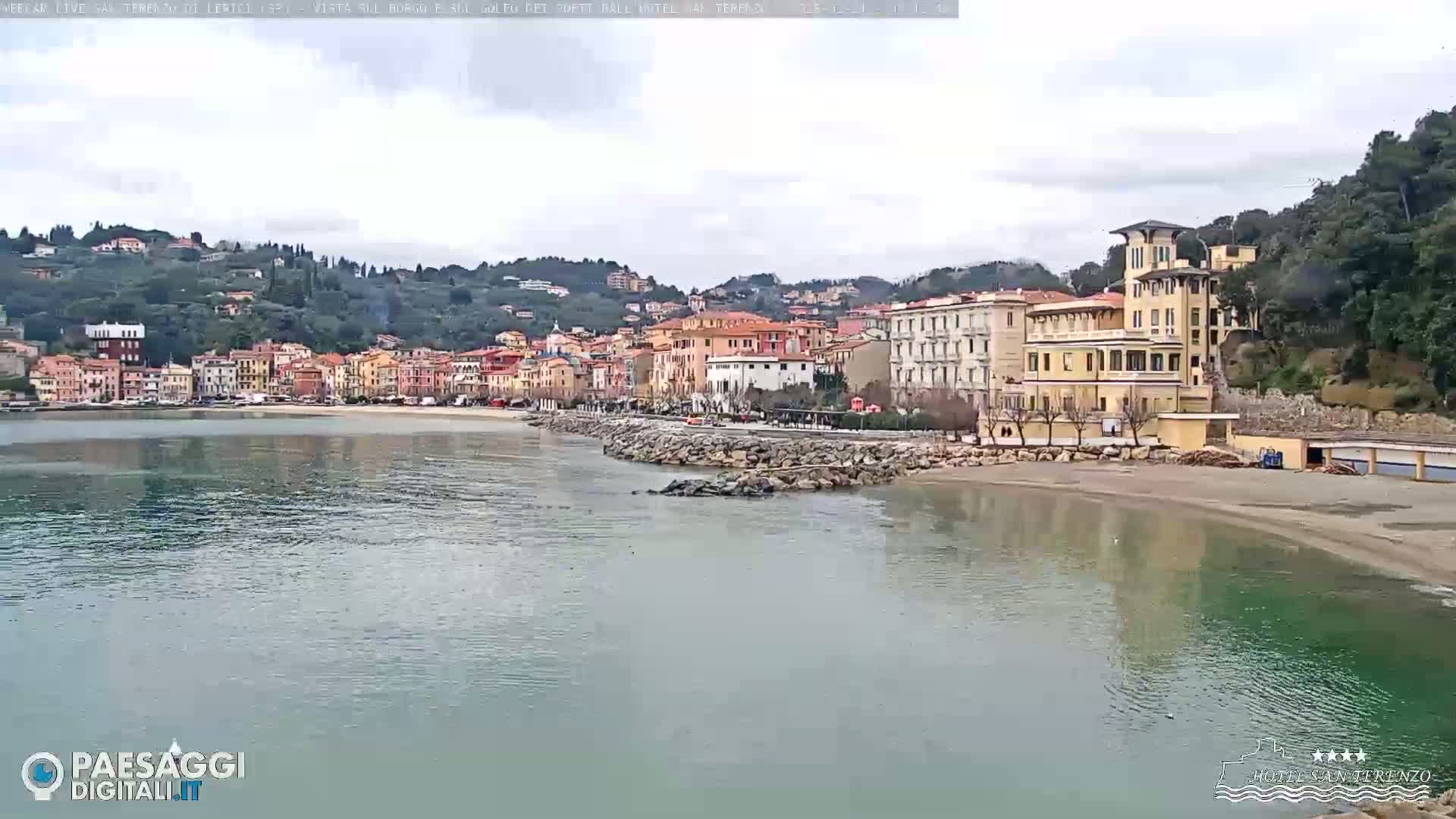 A calm bay with a sandy beach and colorful buildings situated on a hillside under an overcast sky.