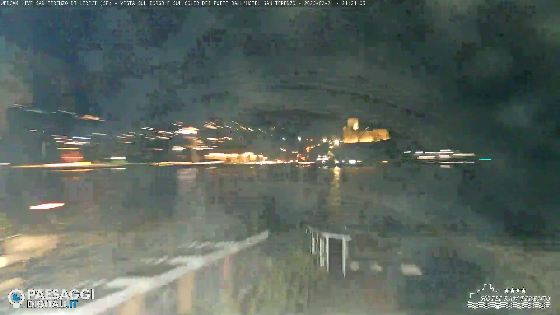 A nighttime view across dark water toward a brightly lit town and a castle, with blurry lights suggesting motion in the foreground.