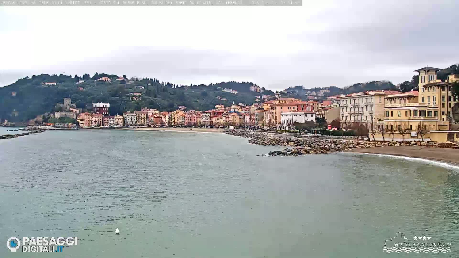 A coastal town with multi-story buildings sits on a gently sloping hill overlooking a calm, gray sea under an overcast sky.
