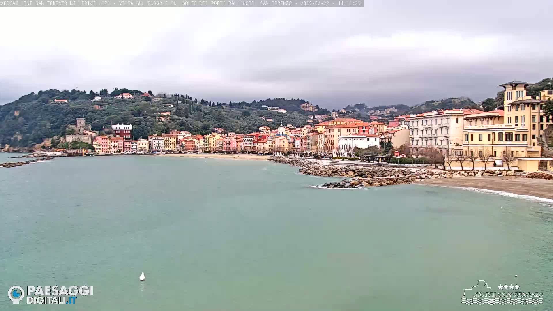 A coastal town with colorful buildings sits on a hillside overlooking a calm, green-blue sea under an overcast sky.