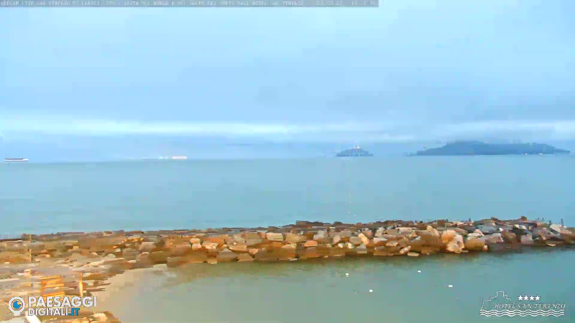 A rocky breakwater sits in calm, light blue water, with hazy islands visible in the distance under an overcast sky.