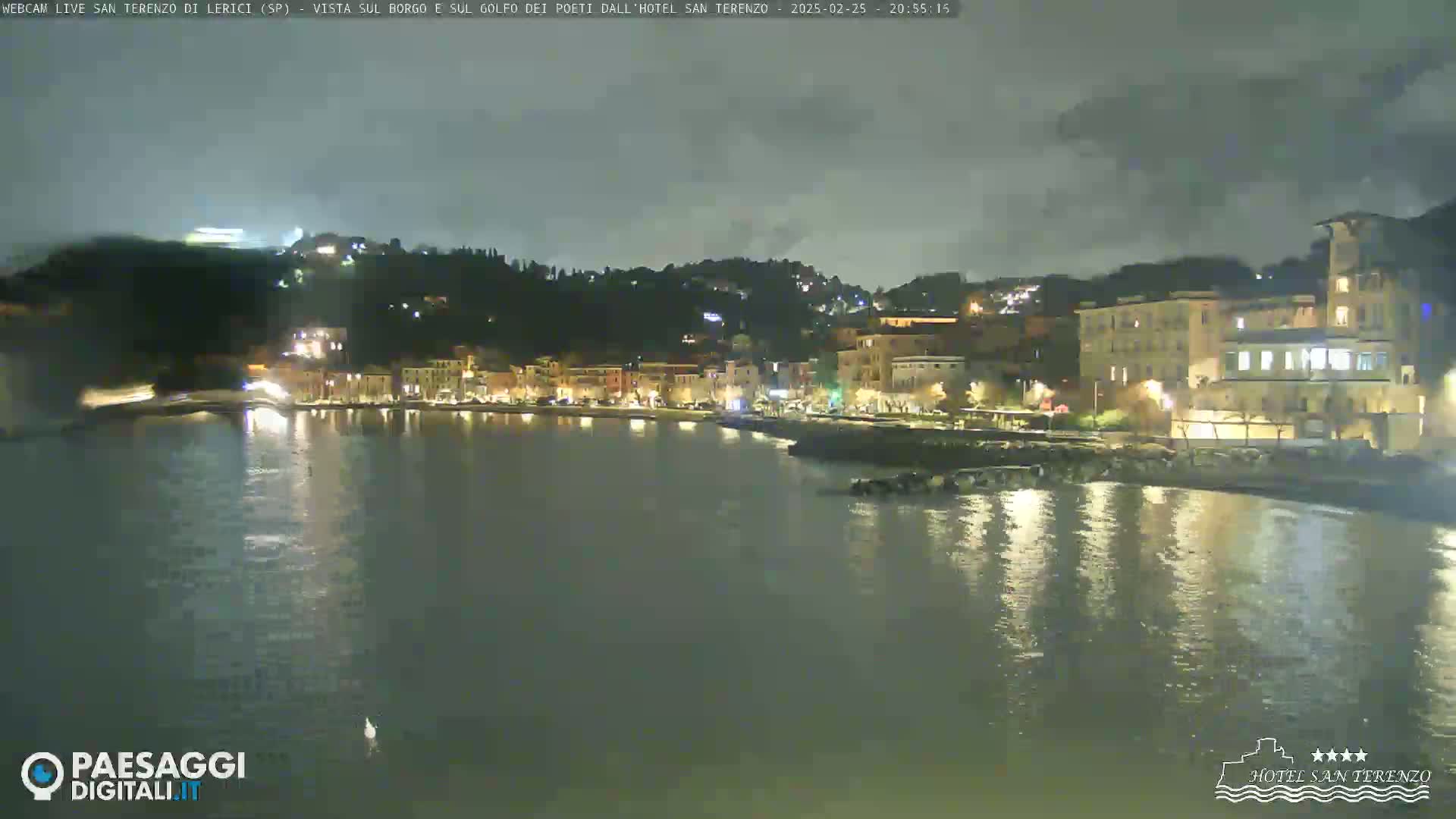 A nighttime view of a coastal town illuminated by lights, reflecting on calm water under a cloudy sky.