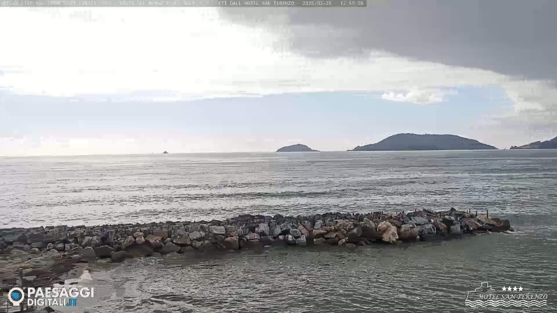 A rocky breakwater extends into a choppy sea under a partly cloudy sky, with islands visible on the horizon.