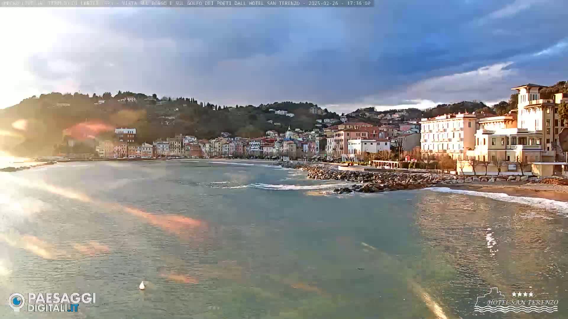 A coastal town with multi-story buildings sits on a hillside overlooking a calm sea under a partly cloudy sky.