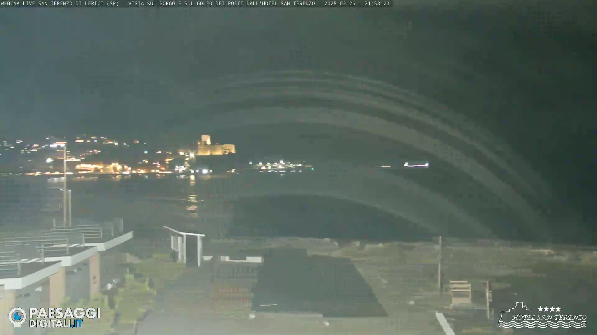 A nighttime view of a dark, calm body of water with a distant, illuminated town and a castle visible across the water.
