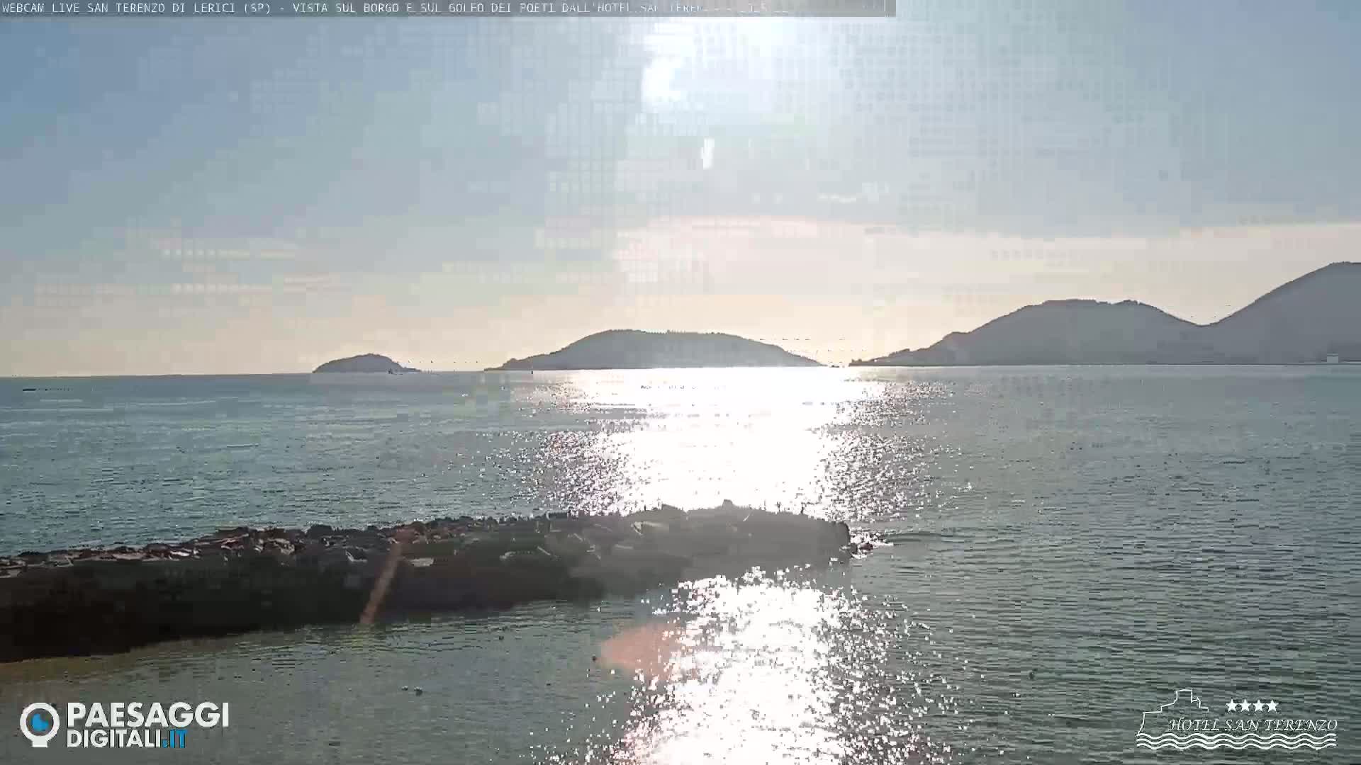 A bright, sunny day reveals a calm sea reflecting sunlight, with distant hills and a rocky outcrop in the foreground.