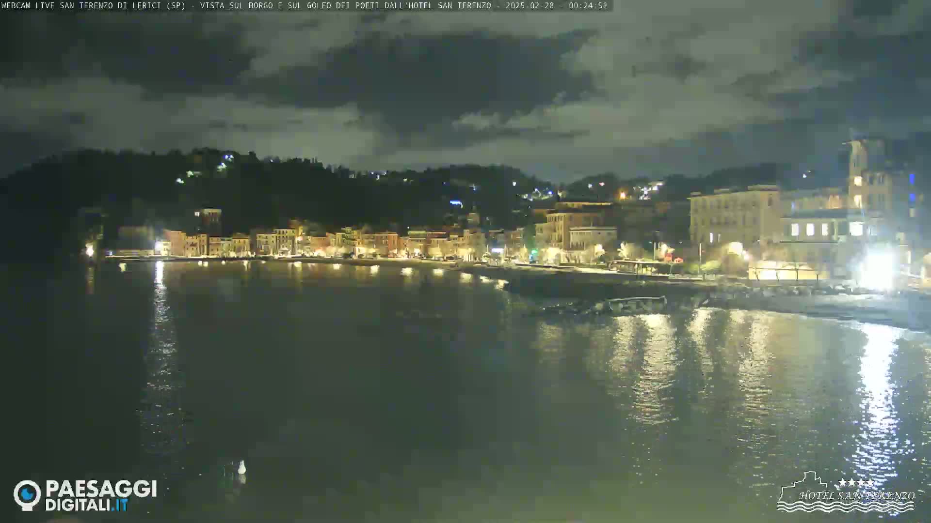A nighttime view of a coastal town with buildings lit up along the waterfront under a partly cloudy sky.