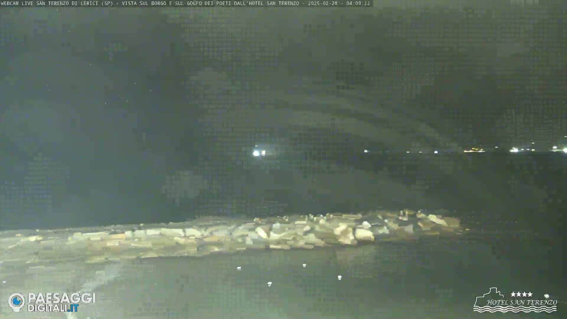 A nighttime view of a rocky shoreline and dark water, with faint lights visible in the distance.