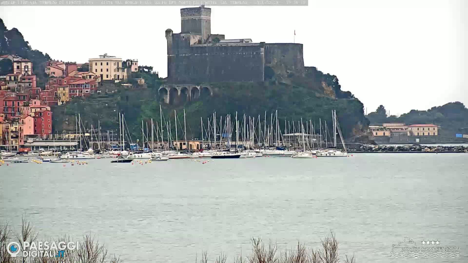 A stone castle sits atop a hill overlooking a harbor filled with sailboats, under an overcast sky.