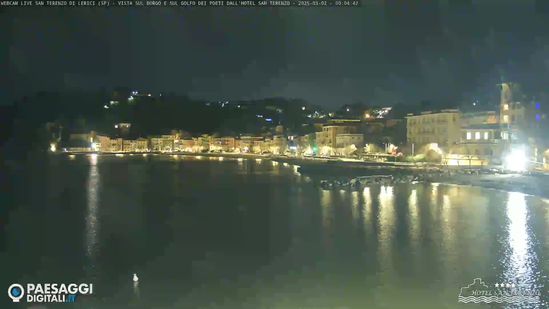A nighttime view of a calm body of water reflecting the lights of a town nestled against a hillside.