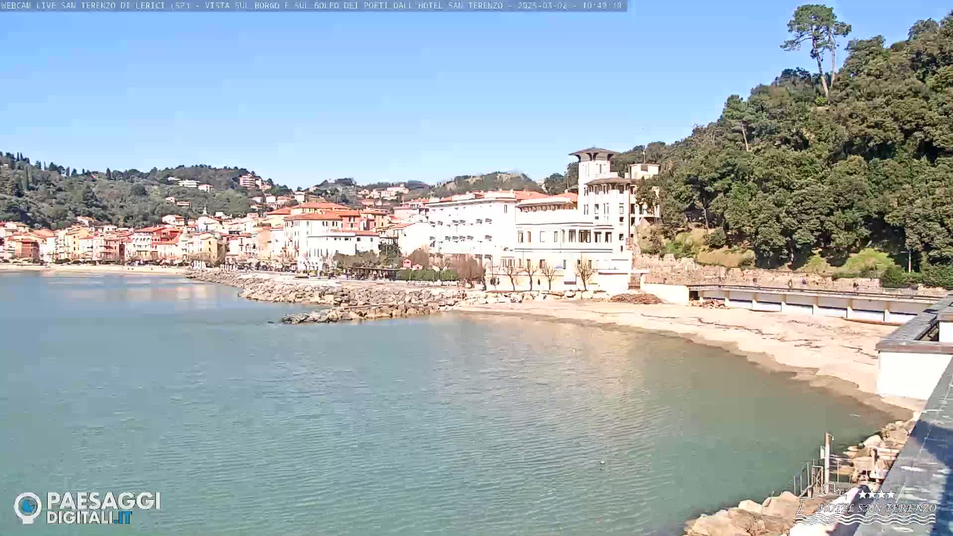 A seaside town on a sunny day with calm water, featuring buildings along the shoreline and hills covered in lush green vegetation in the background.