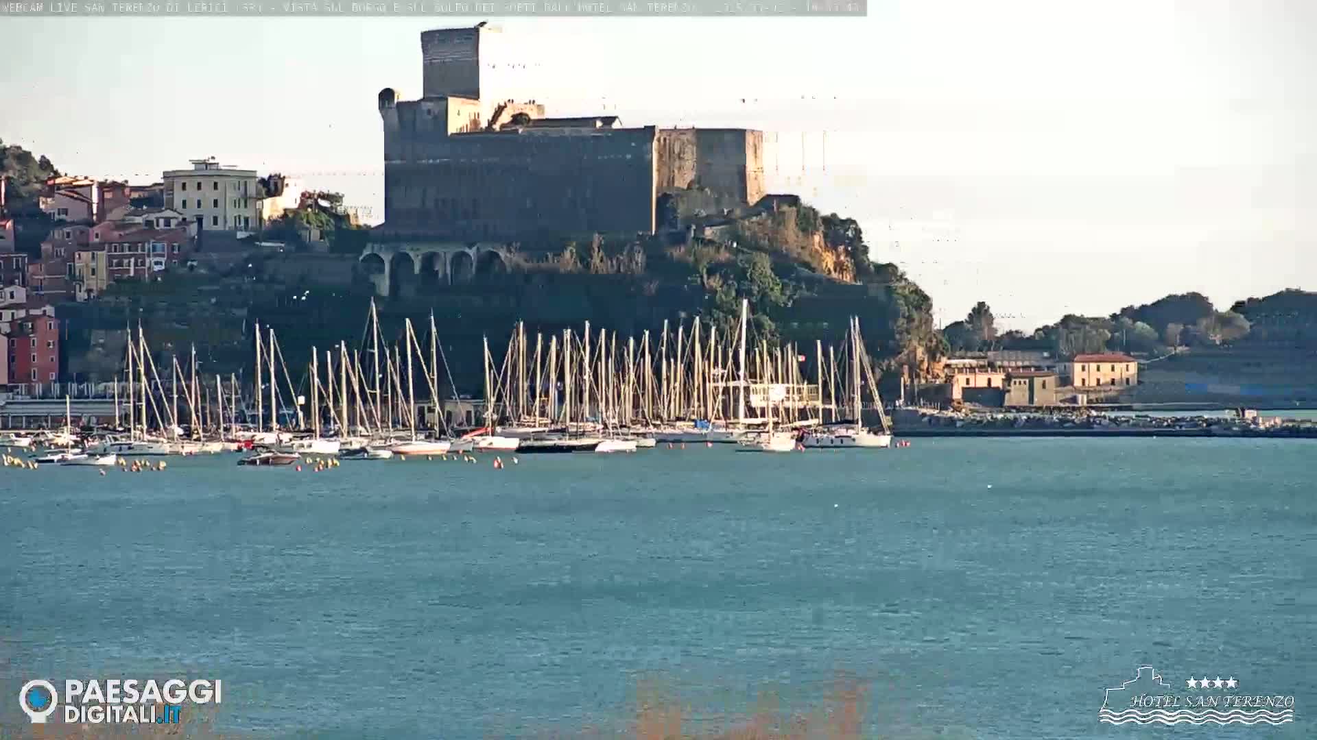 A stone castle sits atop a hill overlooking a harbor filled with sailboats under a mostly clear sky.