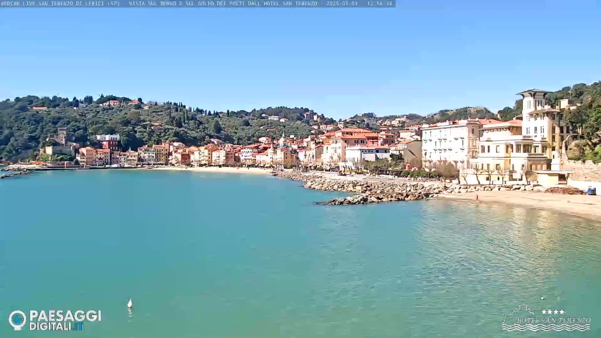 A clear, sunny day reveals a picturesque coastal town nestled against a hillside, with a calm, teal-colored sea lapping at a sandy beach.