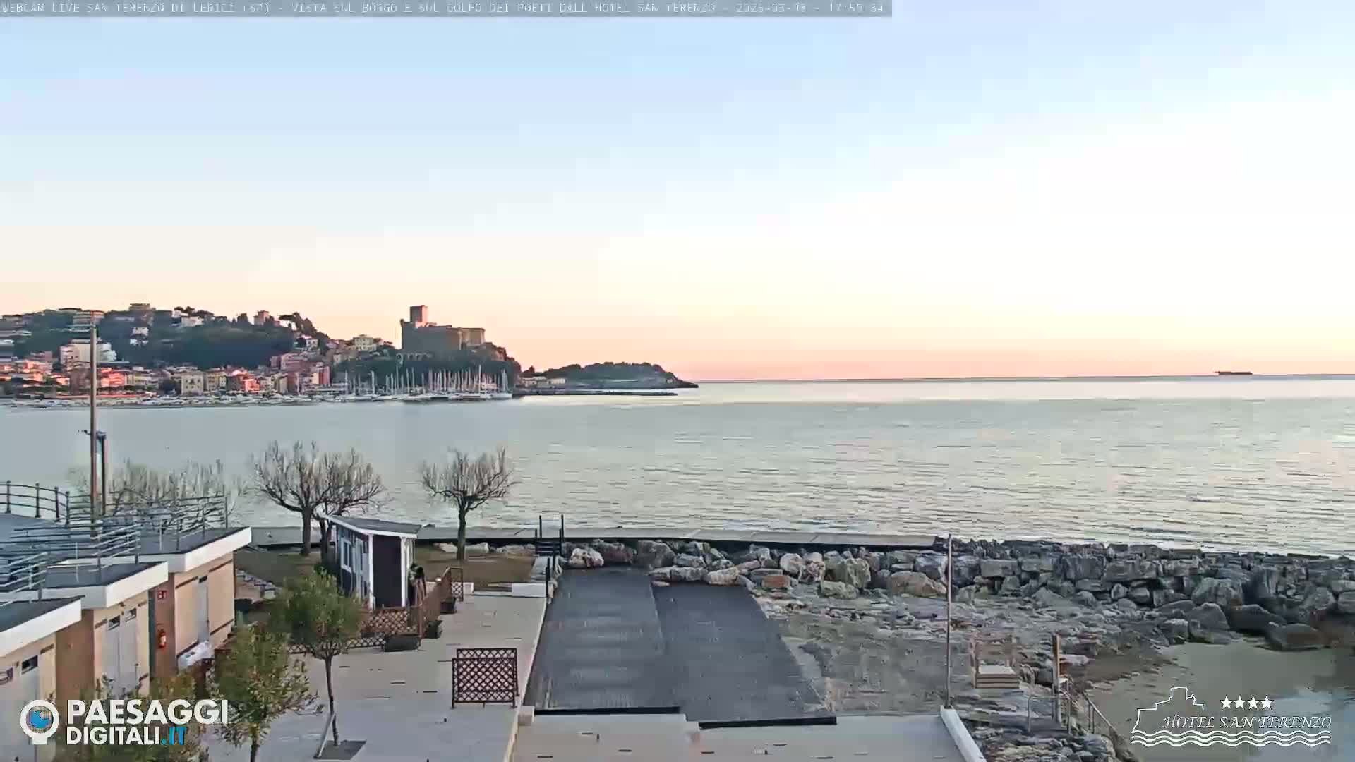A seaside town with a castle on a hill is visible across a calm bay at sunset, with a rocky shore in the foreground.