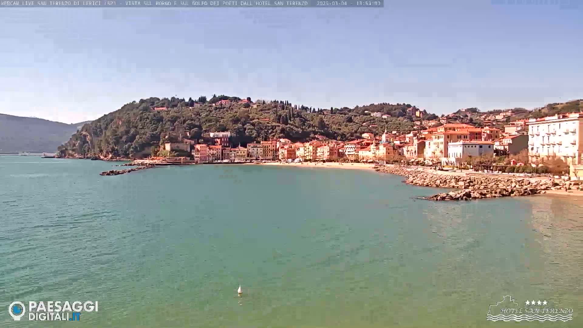 A picturesque coastal town nestled on a hillside overlooking a calm, teal-colored bay under a clear, sunny sky.