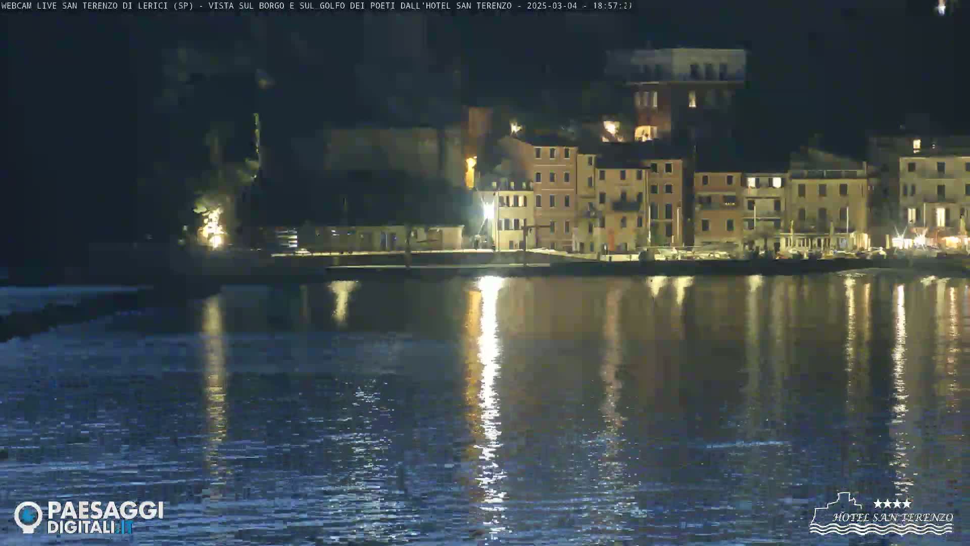 A nighttime view of a waterfront town with multi-story buildings, reflected in calm water.