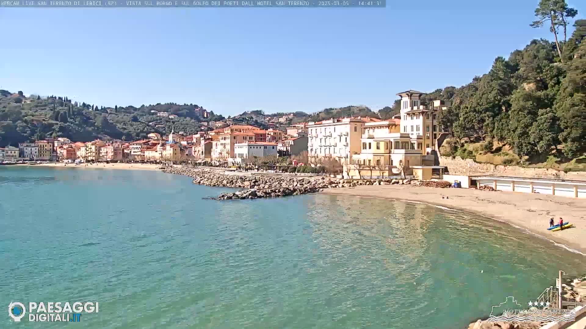 A clear, sunny day reveals a coastal town nestled between hills and a calm, teal sea, with a sandy beach and a few people visible.