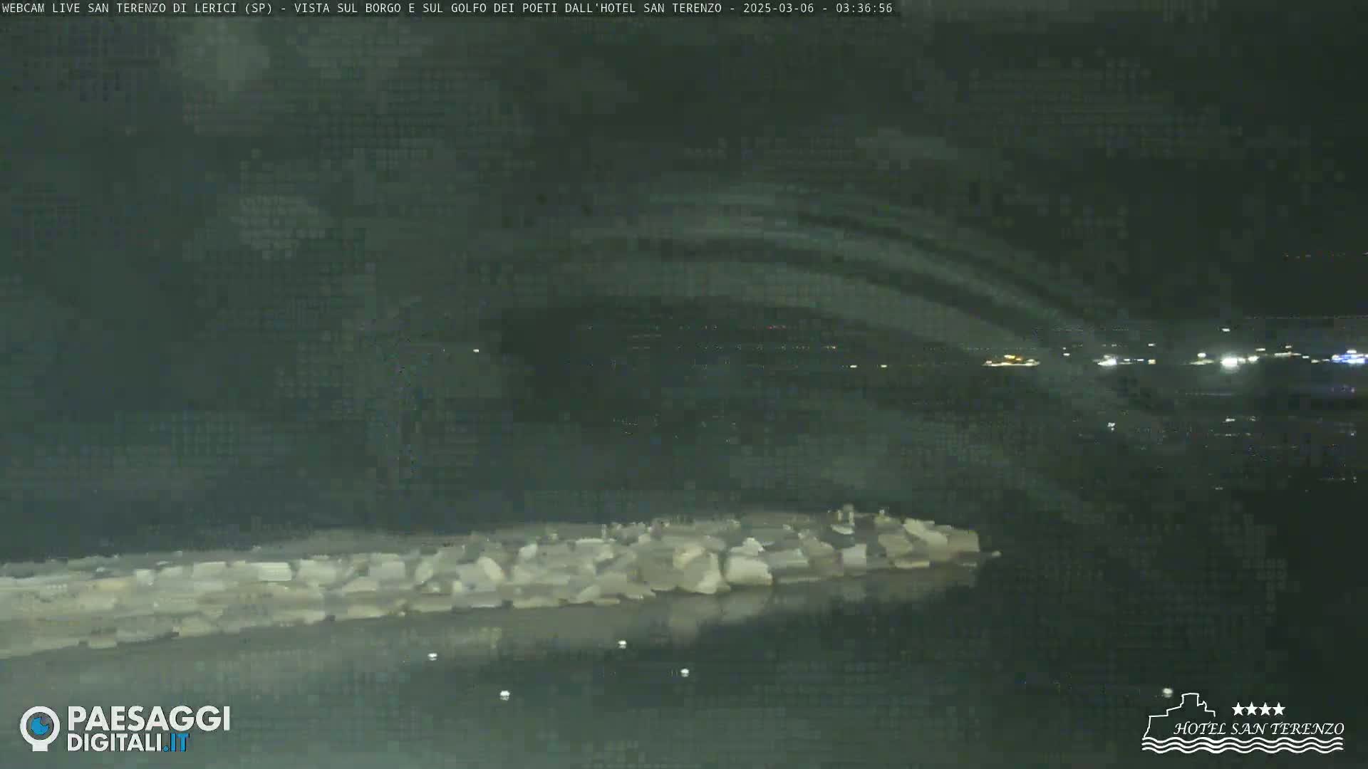 A nighttime, low-light view of a rocky shoreline and calm water, with distant lights visible across the water.
