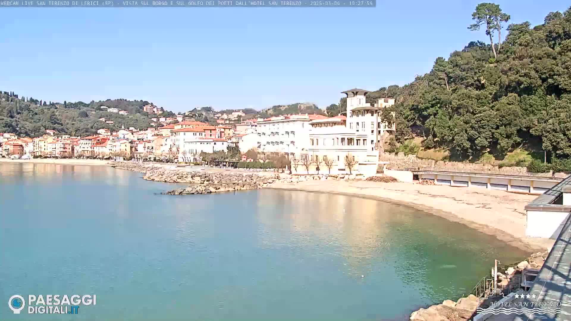 A coastal town with multi-story buildings is situated beside a calm, light-blue bay under a clear, sunny sky.