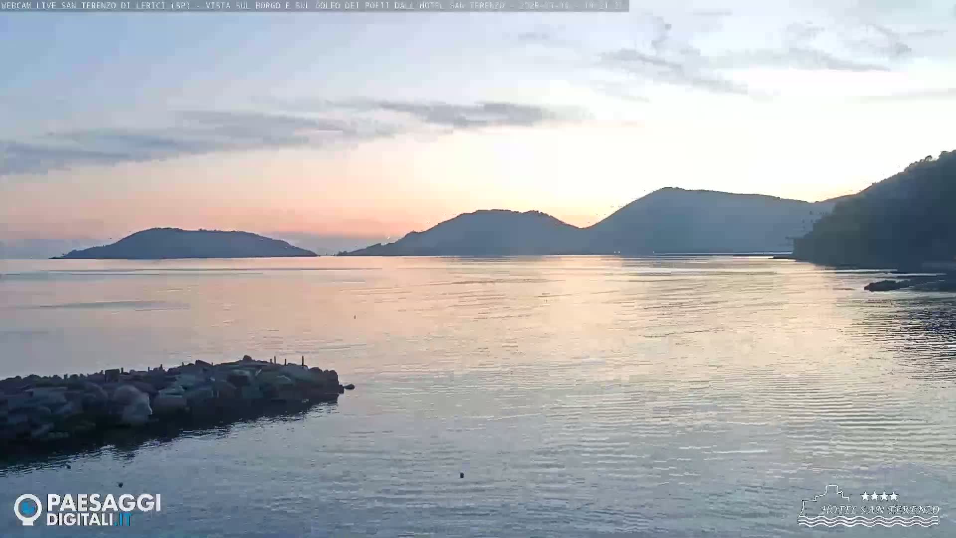 A calm body of water at sunset, with several dark hills and a small rocky jetty in the foreground.