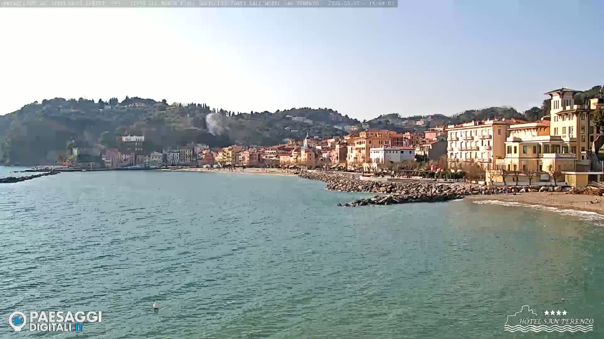 A seaside town nestled against a hillside under a clear, sunny sky, with a calm, blue-green sea stretching out in front.
