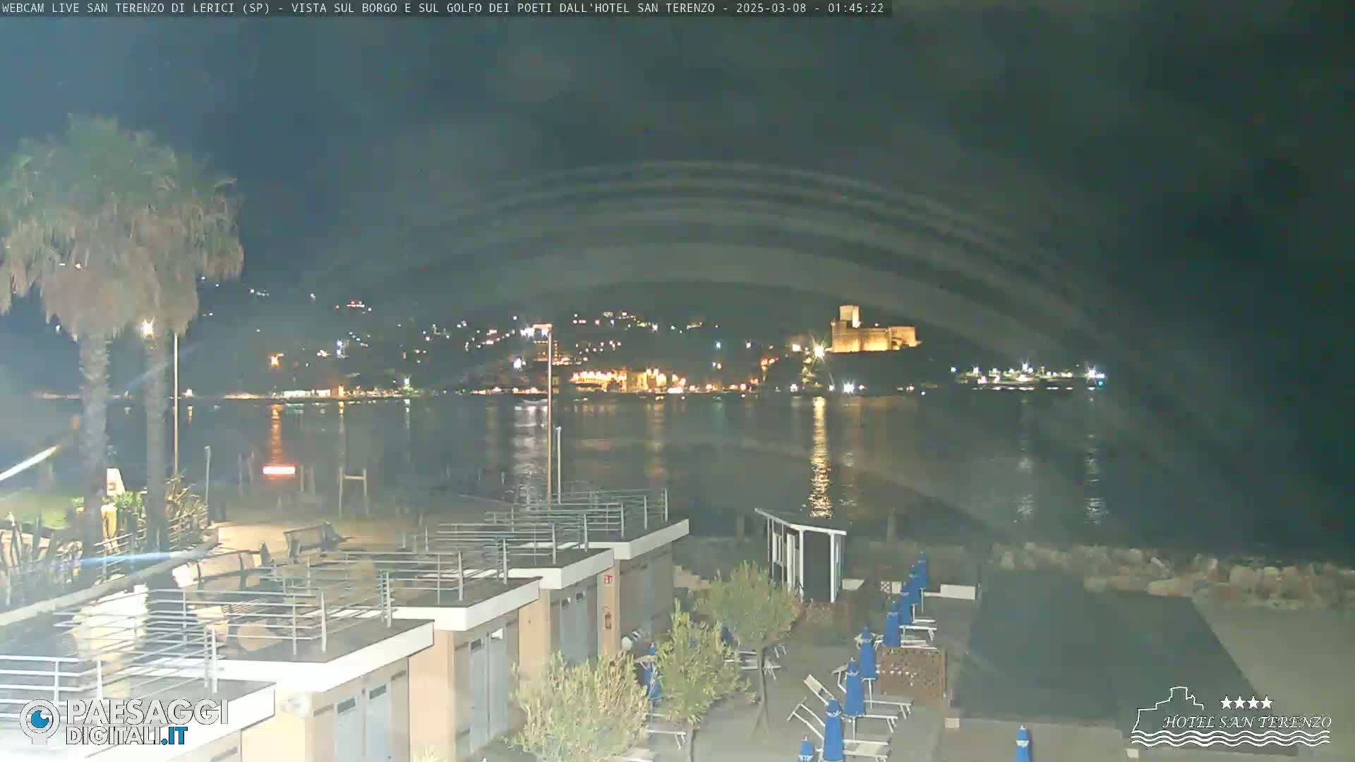 A nighttime view of a calm body of water with a town and castle illuminated on the far shore, seen from a beach with beach huts and lounge chairs.