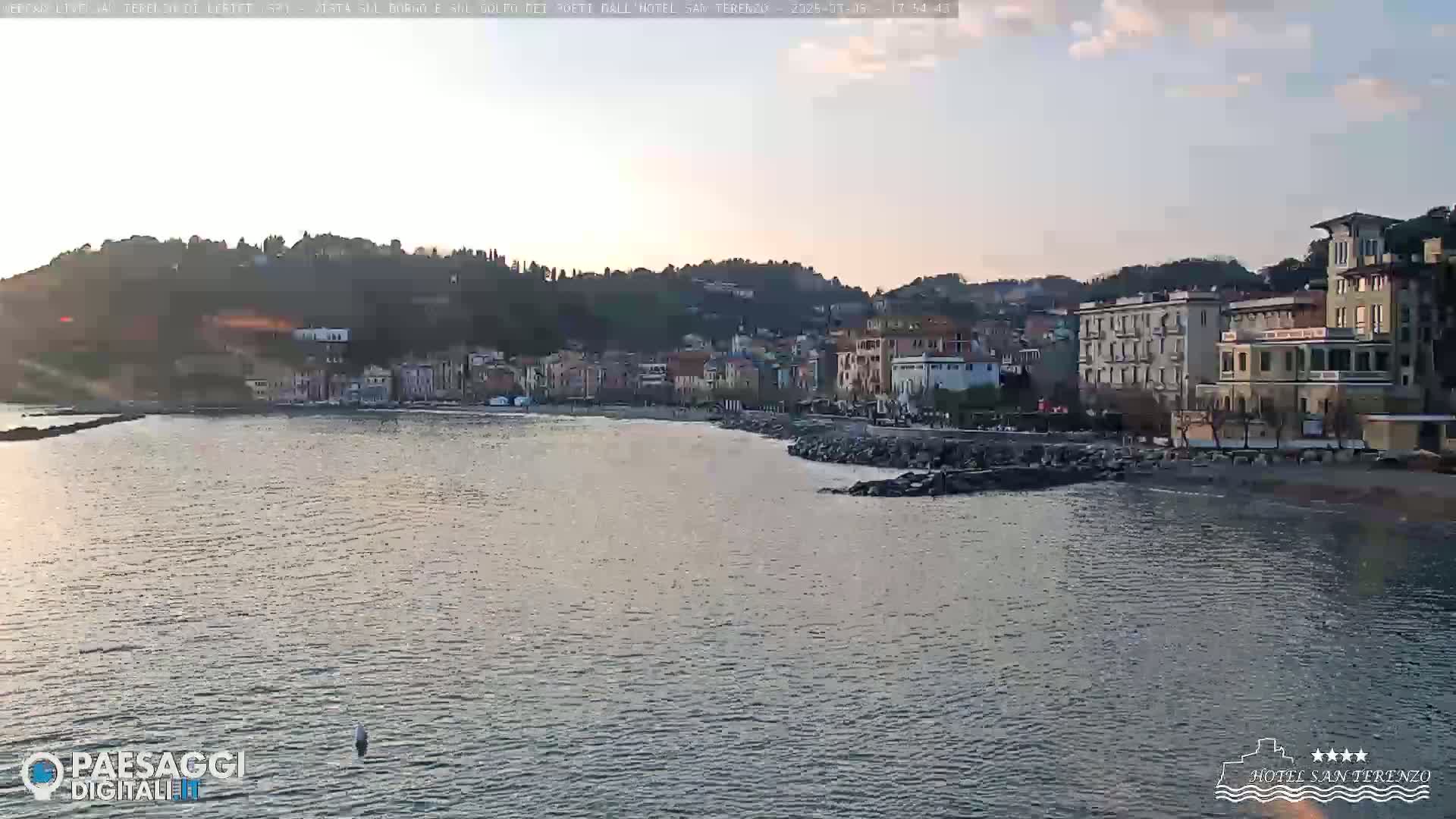 A calm body of water laps against a rocky shore, in front of a town nestled against a hillside under a pale, mostly clear sky.