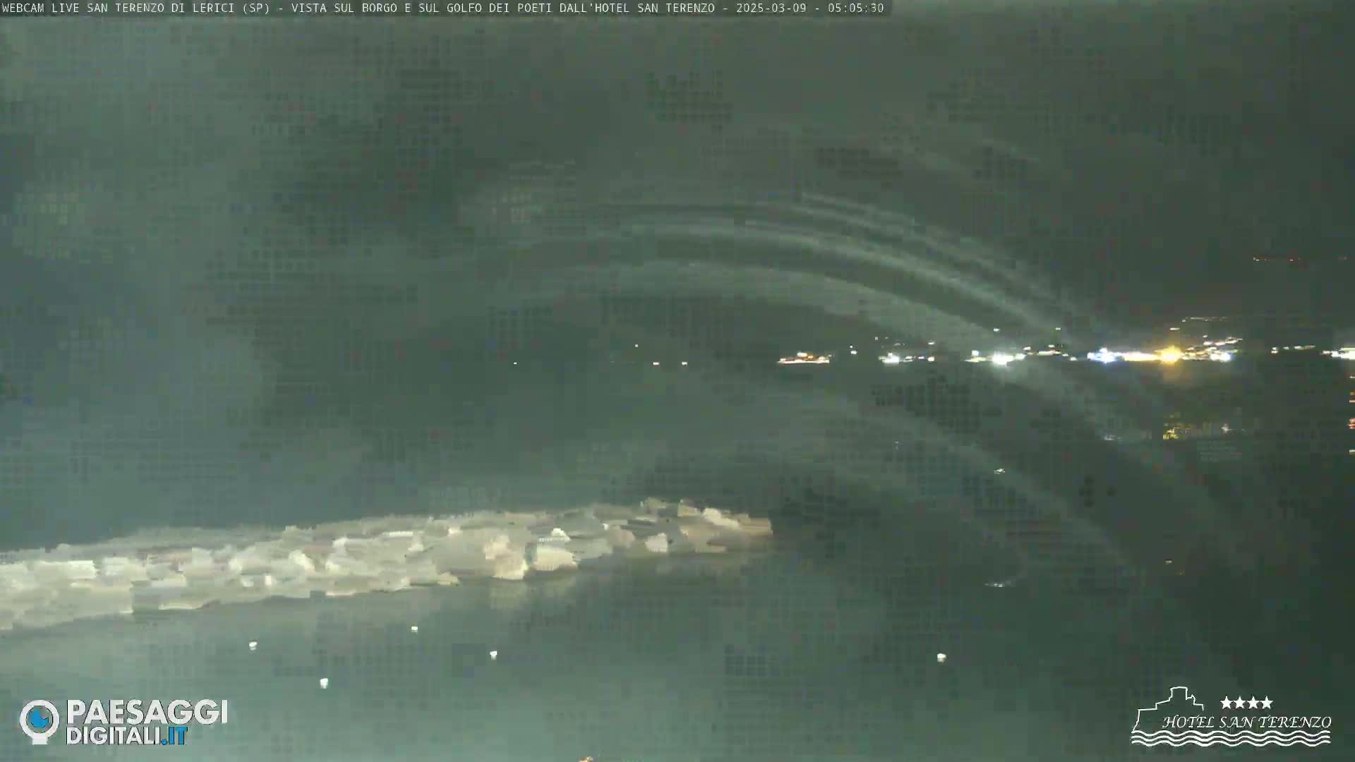 A nighttime, long shot of a dark body of water with a shoreline in the foreground, and lights from a town visible in the distance under a mostly clear sky.