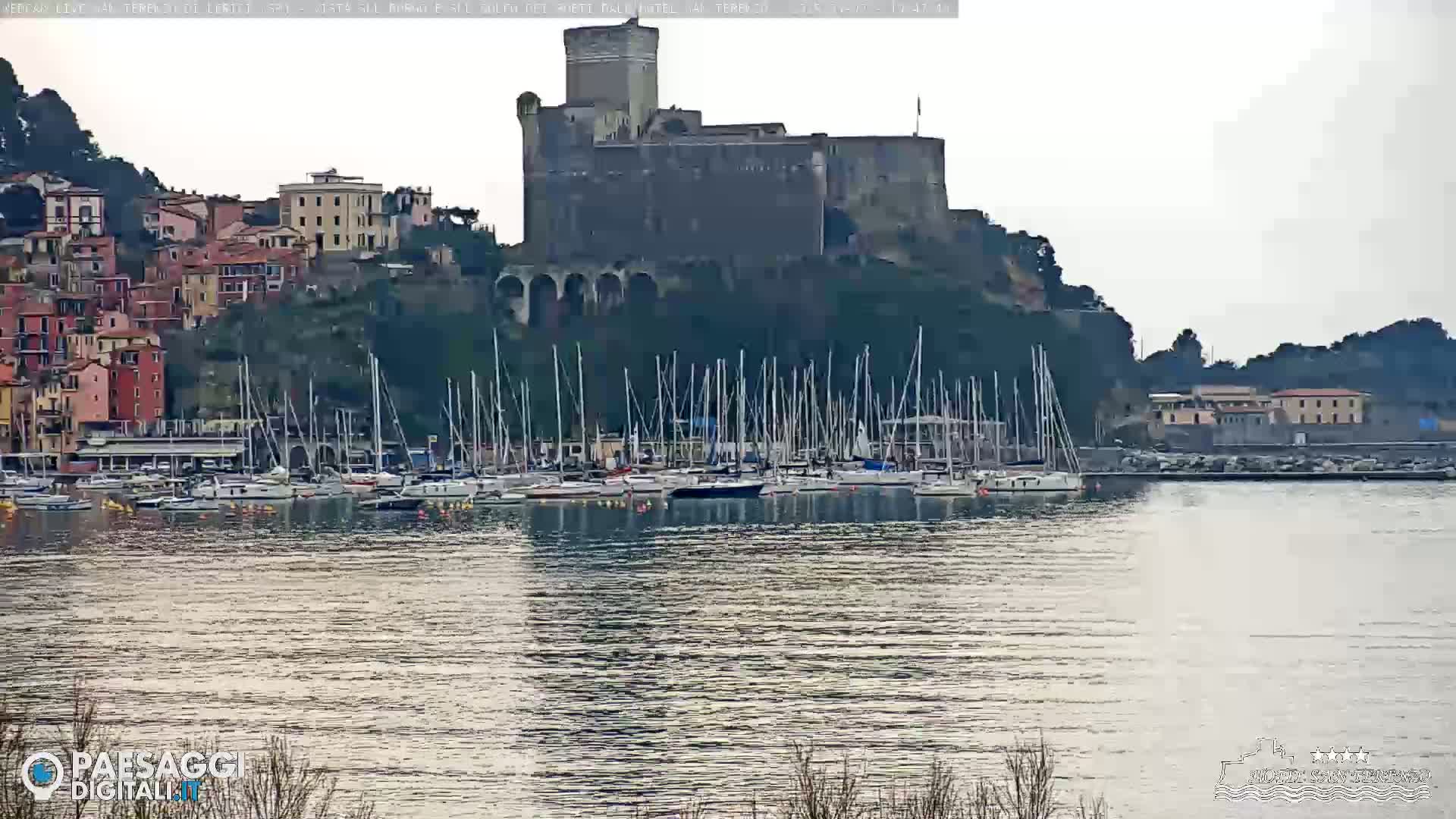 A stone castle sits atop a hill overlooking a harbor filled with sailboats, under a cloudy sky.