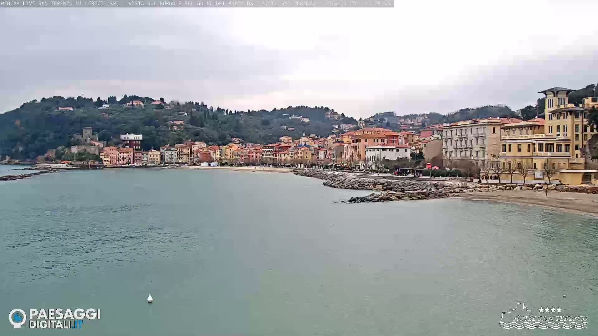 A coastal town with multi-story buildings sits on a hillside overlooking a calm bay under an overcast sky.