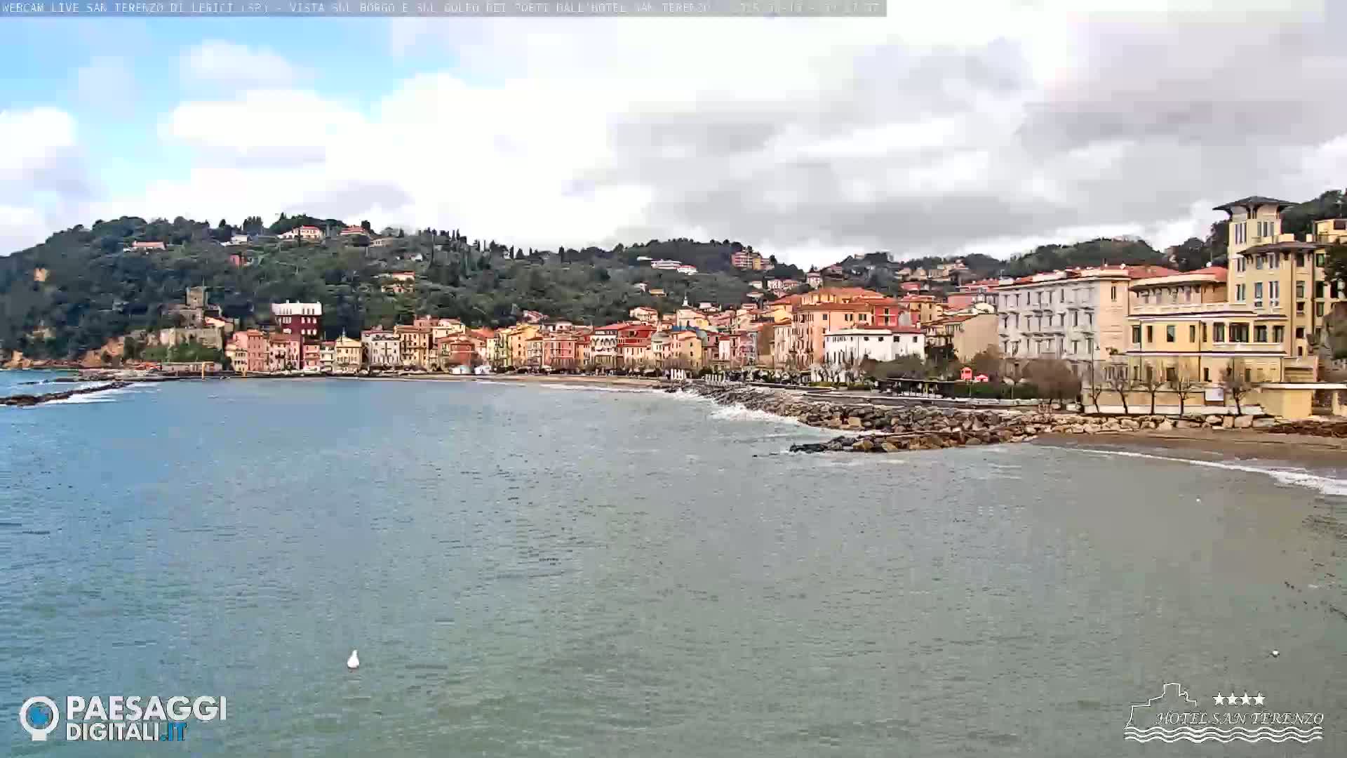 A coastal town with multi-story buildings sits on a hillside overlooking a calm sea under a cloudy sky.