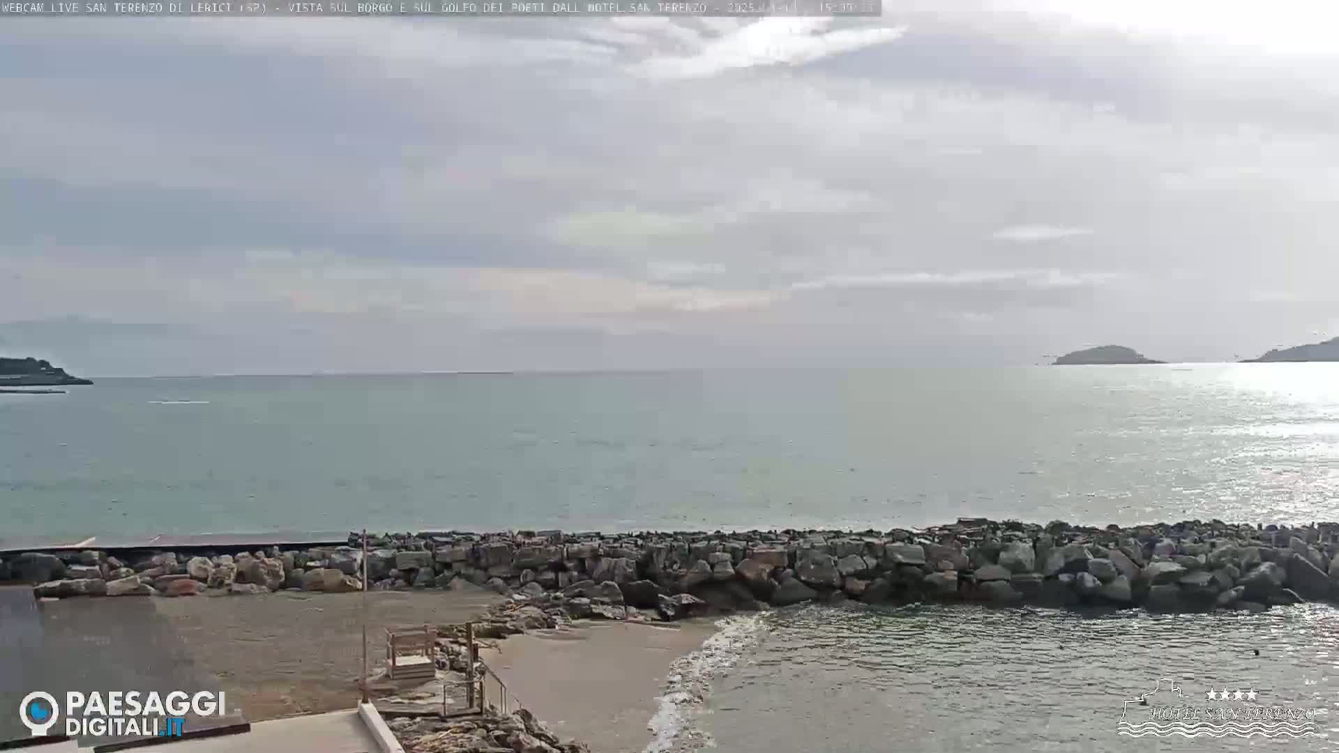 A rocky breakwater protects a small sandy beach from a calm, light-grey sea under an overcast sky.