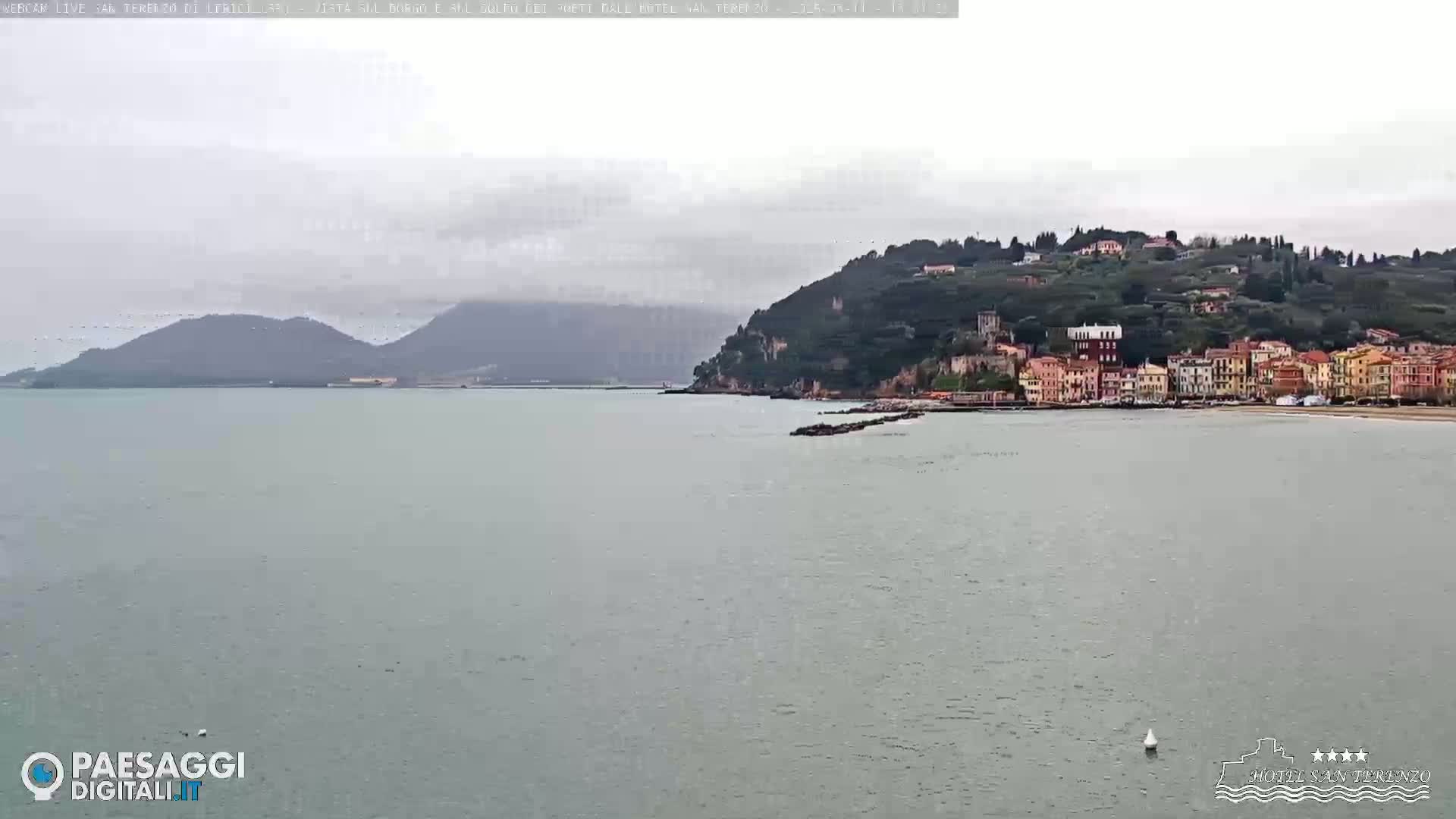 A calm sea stretches out before a coastal town nestled against a verdant hillside under an overcast sky.