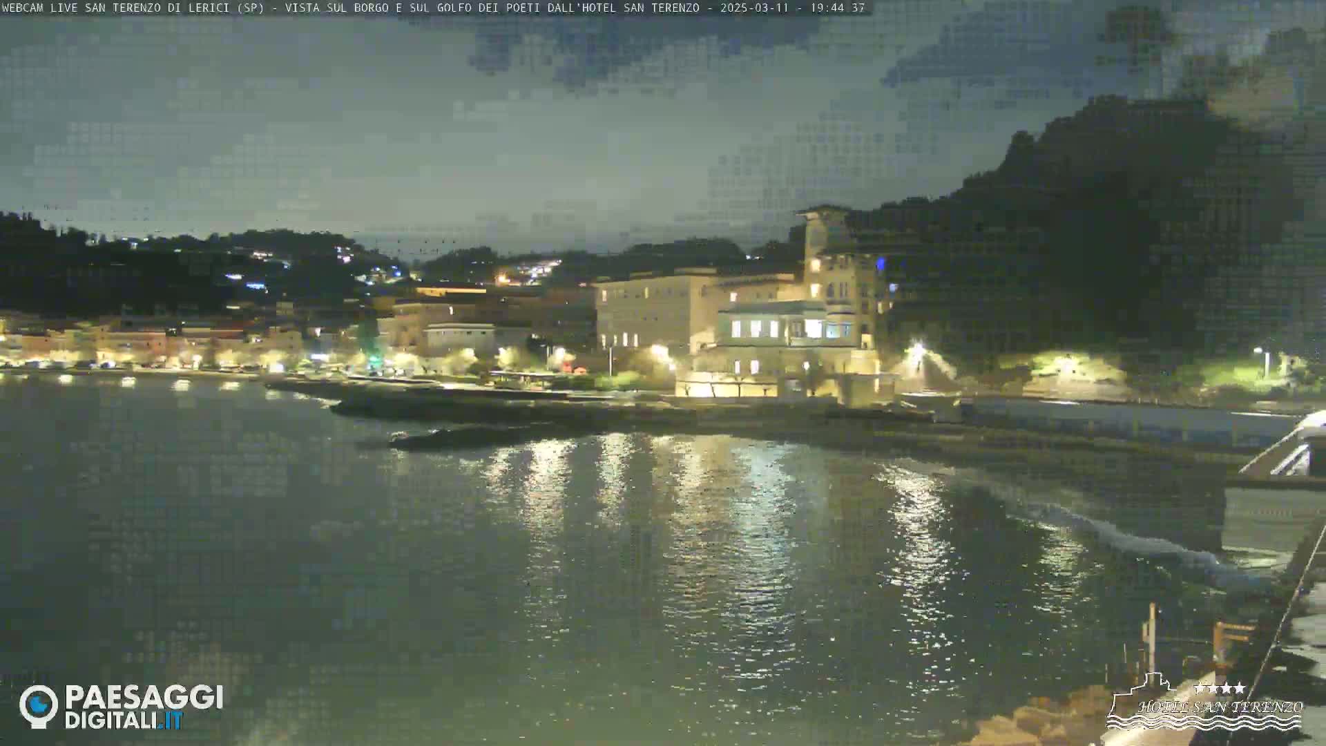 A nighttime view of a coastal town with buildings illuminated along the water's edge, under a partly cloudy sky.