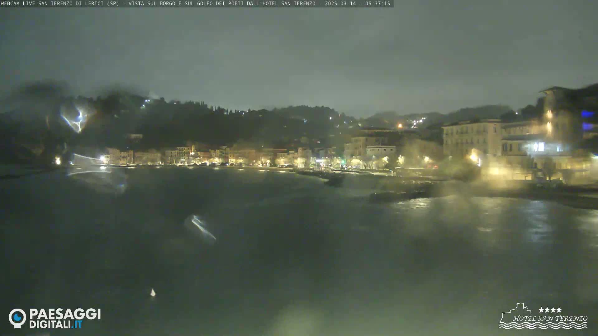 A nighttime view of a rain-swept coastal town with illuminated buildings along a dark, choppy waterfront.