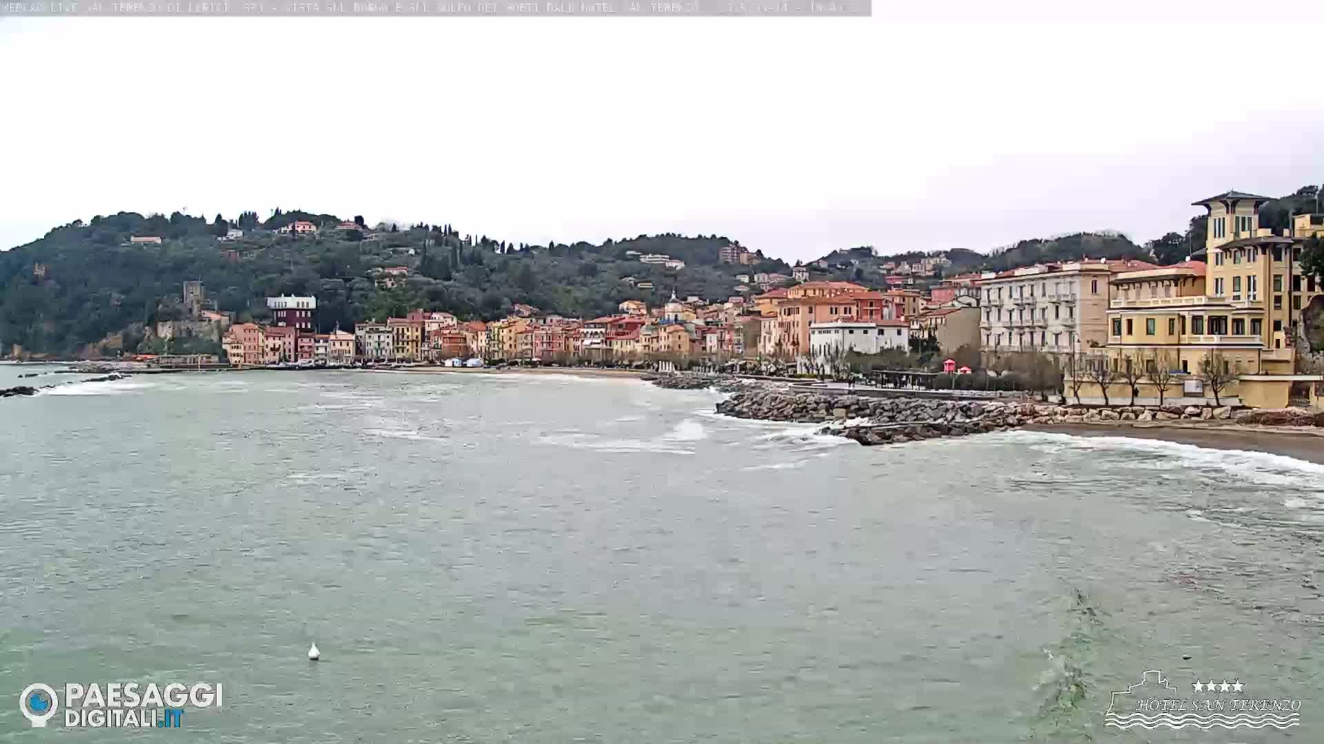 A coastal town with multi-story buildings sits on a hill overlooking a calm sea with small waves on an overcast day.